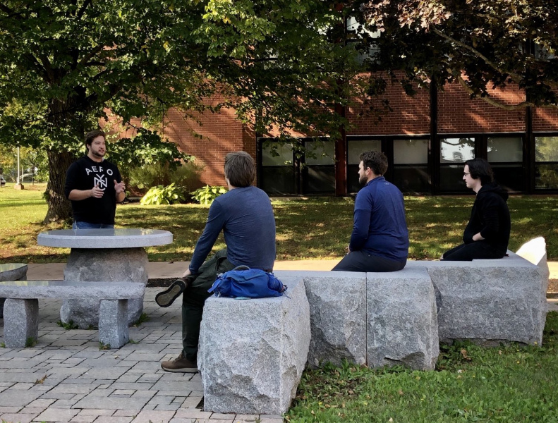 Three students sit on stone slabs in an outdoor setting as they listen to a fourth student instructing them