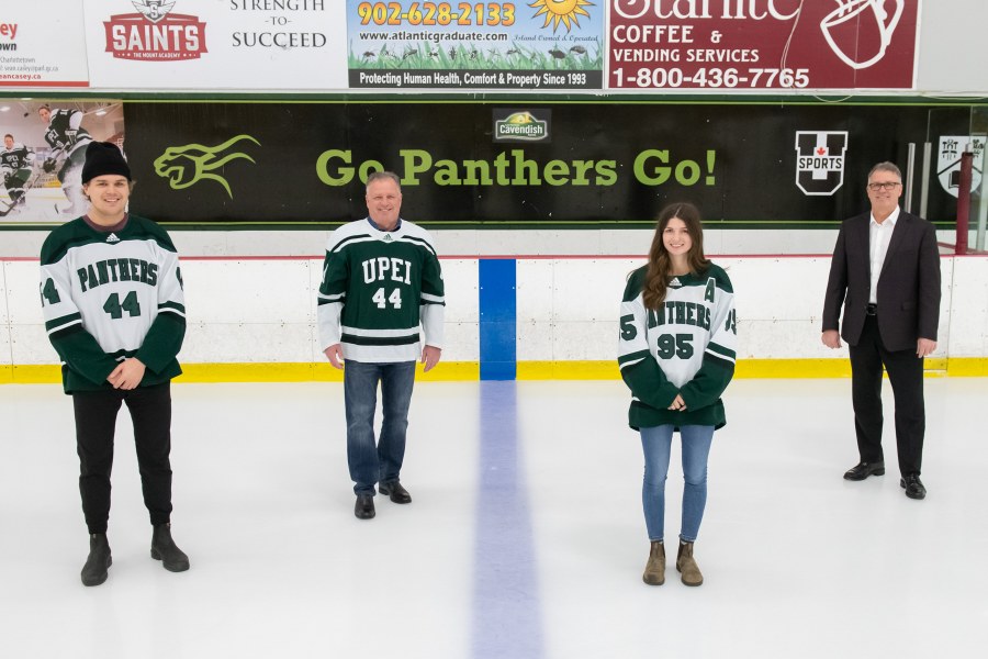 Four people stand on a skating rink surface. A young male hockey player, an older man, a young female hockey player, and another older man