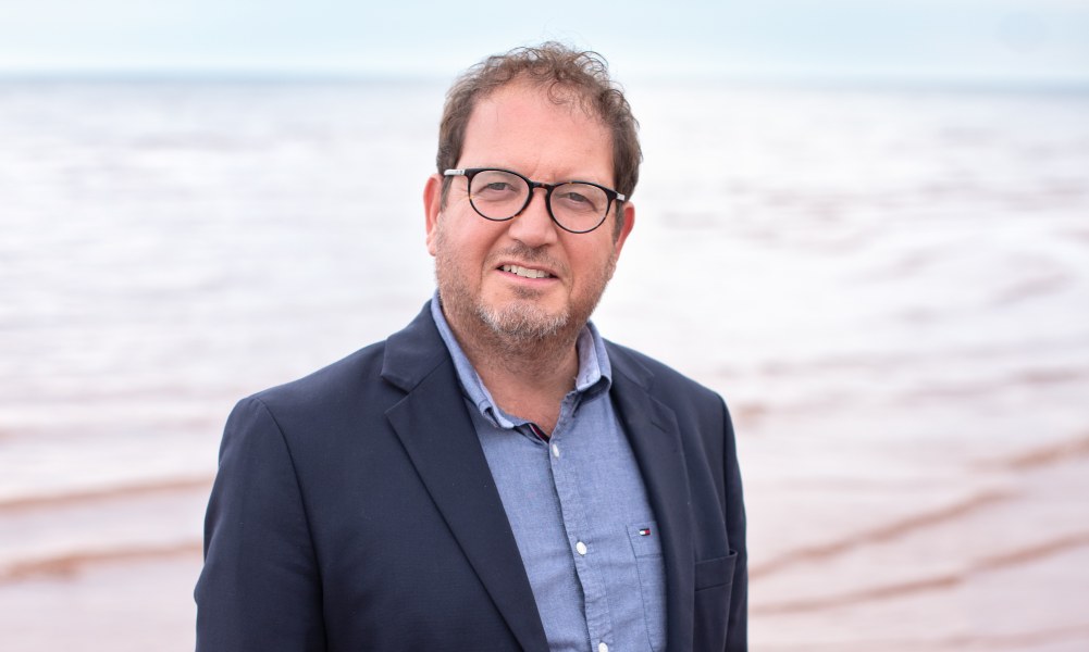 A smiling man in a jacket and unbuttoned shirt. Photo taken at the beach.