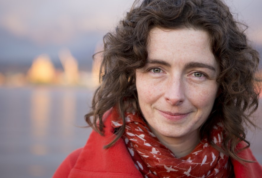 Bren Simmers A photo portrait of a smiling woman with medium length dark curly hair. She is standing in front of a shoreline with water in the background.