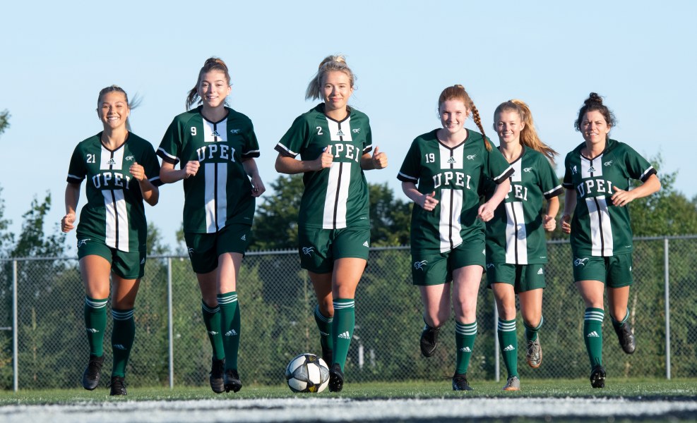 A squad of female soccer players runs towards the camera across a soccer pitch