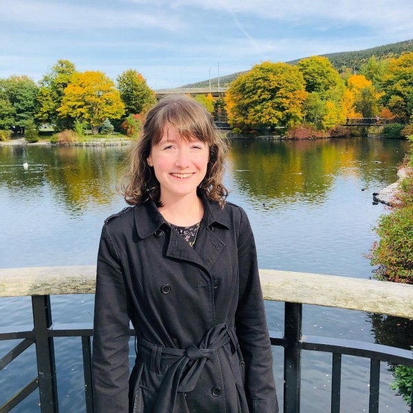 A smiling young woman stands in front of a beautiful scene with a lake and trees