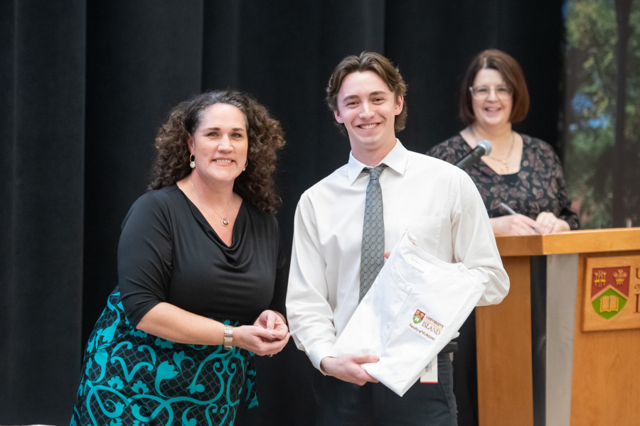 UPEI Faculty of Nursing Interim Dean, Dr. Christina Murray presents nursing student Scott Davis with a scrub coat as Interim Associate Dean, Patrice Drake looks on