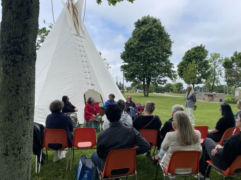 Group of people listening to Indigenous speakers, sitting in front of a tipi