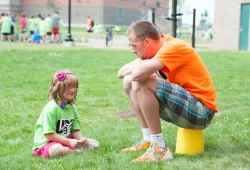 A young girl sits on the grass speaking with an adult