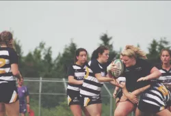 A woman in a black uniform holding a rugby ball charges through a group of women wearing striped uniforms