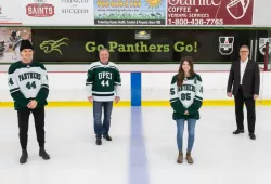 Four people stand on a skating rink surface. A young male hockey player, an older man, a young female hockey player, and another older man