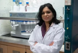 A female scientist in a white lab coat stands with her arms crossed in front of a laboratory environment in which complicated-looking scientific formulae are written out on a glass wall