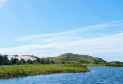 A grassy sand dune overlooking deep blue water and a sunny sky