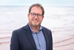 A smiling man in a jacket and unbuttoned shirt. Photo taken at the beach.