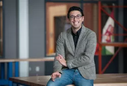 A smiling man in a jacket and unbuttoned shirt. He sits casually on the corner of a workbench in an engineering lab.