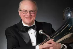 A smiling man in a black tuxedo cradles a tuba in his arms