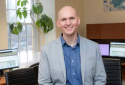 A smiling man in a jacket and unbuttoned shirt. He is sitting in an office with natural light and office plants