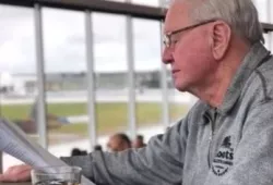 Image of Billy Mulligan seated at a table with a beverage reading a harness racing program 