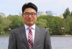 A smartly dressed man in a grey suit stands in front of a river with distant buildings in the background