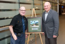 Two men standing next a framed photo of AVC