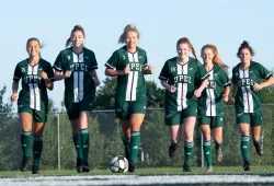 A squad of female soccer players runs towards the camera across a soccer pitch