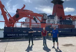 Three smiling men stand on a wharf next to a giant ship festooned with cranes and other research material