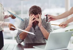 Man sitting at desk holding his head in his hands surrounded by stressors