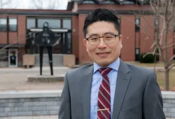 photo of man wearing glasses standing in front of a metal sculpture and red brick building