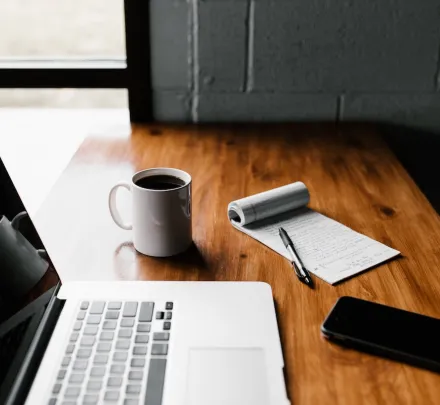 wooden table with a laptop computer, cup of coffee, cellular phone, and notepad