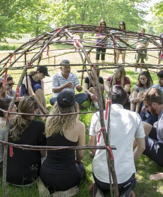 an indigenous elder and a group of people sitting inside the wooden frame of a sweat lodge